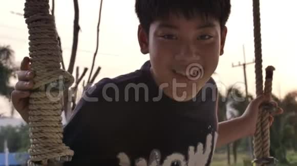 亚洲儿童男孩在家玩秋千与日落耀斑的肖像手持慢动作青少年在秋千上微笑的脸视频的预览图