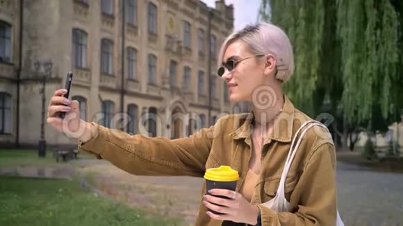 金发时尚的女人戴着太阳镜自拍和拿着咖啡站在大学附近的公园里视频的预览图