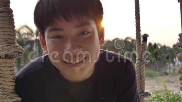亚洲儿童男孩在家玩秋千与日落耀斑的肖像手持慢动作青少年在秋千上微笑的脸视频的预览图
