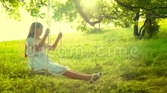 美丽的女孩一头长发穿着白色连衣裙在灿烂的金色夕阳中荡秋千在夏日的橡树下微笑视频的预览图