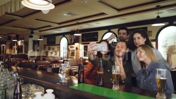 朋友们正在酒吧里用智能手机自拍年轻人摆姿势笑着说话啤酒瓶和视频的预览图