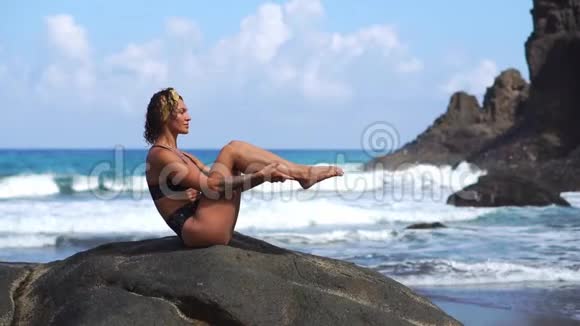 一个穿比基尼的年轻女孩坐在石头上交替地举起腹部肌肉做运动视频的预览图