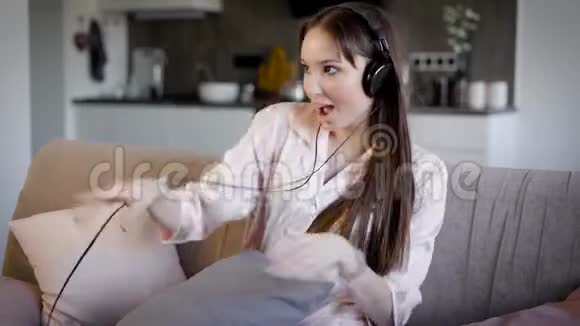 年轻女子坐在沙发上的客厅里带着耳机听音乐她唱歌玩得很开心视频的预览图