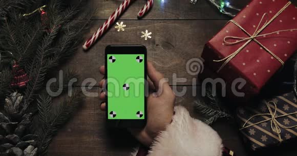 圣诞老人的手在黑色智能手机上用绿色屏幕敲击页面另一只手放一些礼物盒视频的预览图