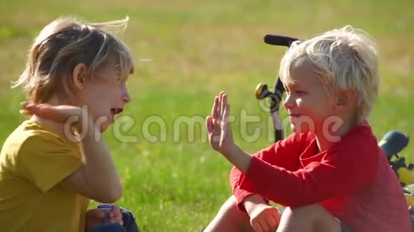 两个小男孩骑自行车的慢镜头坐在草坪上给对方五个成功的概念友谊视频的预览图