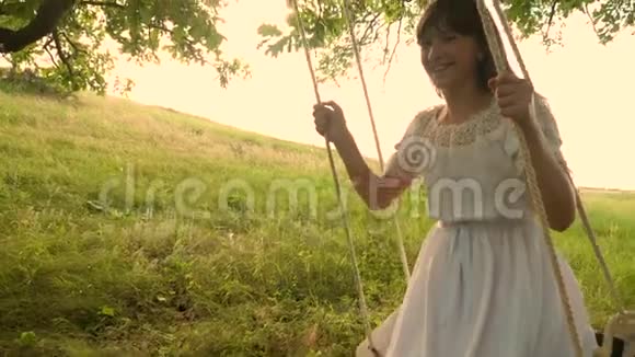 在一个温暖的季节里一个留着白色连衣裙的长发的少女在夏天橡树下的秋千上笑着滚动慢慢视频的预览图
