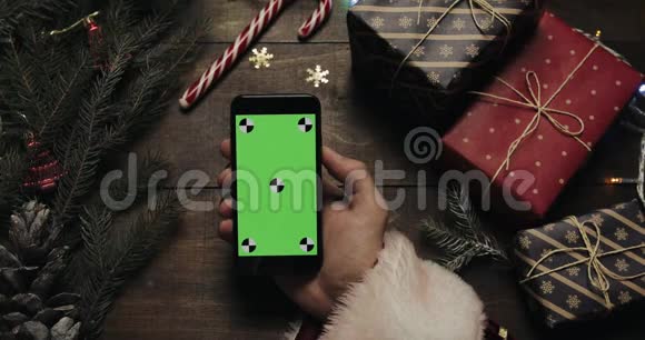 圣诞老人手里拿着绿色屏幕的黑色智能手机另一只手在桌子上放一些礼物盒视频的预览图