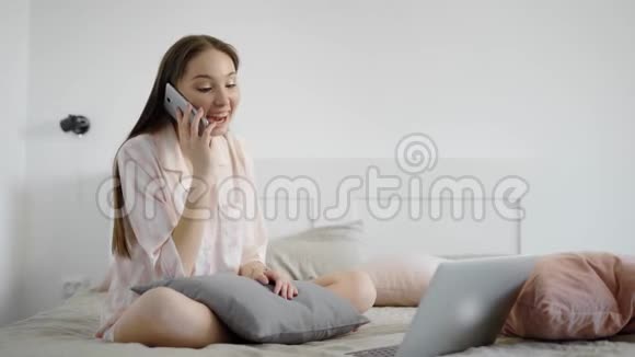 年轻的女人带着笔记本电脑在卧室里女士早上用手机说话可爱的微笑着视频的预览图