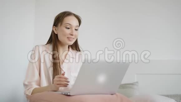 年轻可爱的女人在社交网络上和朋友聊天她双脚在卧室里拿着笔记本电脑视频的预览图