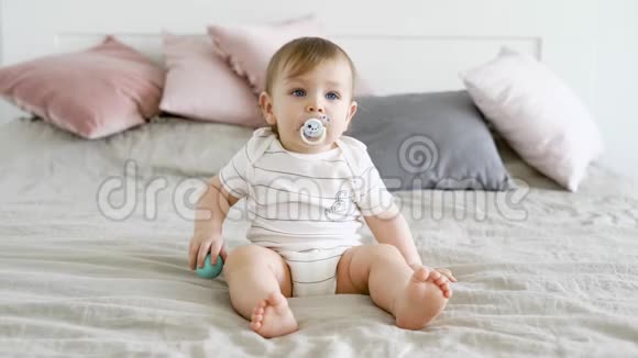 一个可爱有趣的孩子坐在床上嘴里放着奶嘴手里拿着球男孩看着前方视频的预览图