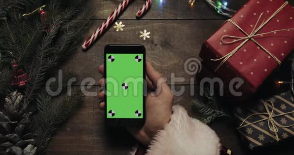 圣诞老人手里拿着绿色屏幕的黑色智能手机另一只手在桌子上放一些礼物盒视频的预览图