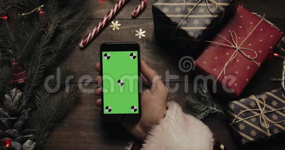 圣诞老人的手在黑色智能手机上用绿色屏幕敲击页面另一只手放一些礼物盒视频的预览图