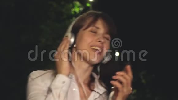 女孩戴着耳机和触摸电话挥舞着长发唱歌跳舞在公园的夜晚音乐中微笑和凯福亚视频的预览图