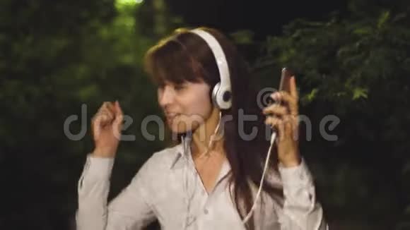 女孩戴着耳机和触摸电话挥舞着长发唱歌跳舞在公园的夜晚音乐中微笑和凯福亚视频的预览图