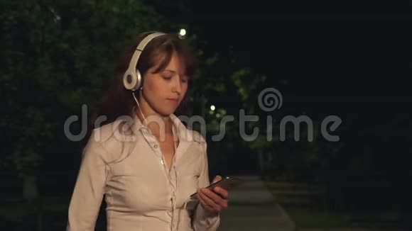 女孩戴着耳机和触摸电话挥舞着长发微笑着跳舞在公园的夜晚从音乐中得到嗡嗡声视频的预览图
