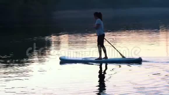 长镜头美丽的女孩漂浮站在苏台上在日落时由桨划着视频的预览图