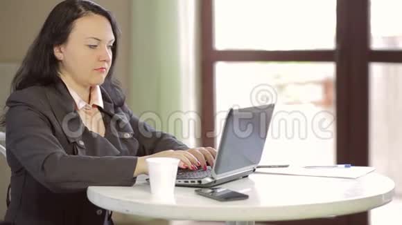 年轻的女人在咖啡馆的一张桌子上在笔记本电脑后面工作喝咖啡慢点视频的预览图