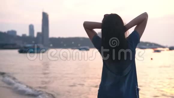 穿着夏装的年轻女子站在热带城市的海滩上日落时分望着大海视频的预览图