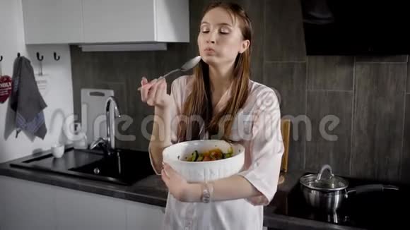 年轻女子正用勺子吃蔬菜沙拉手里拿着大白沙拉碗晚上站在厨房里视频的预览图