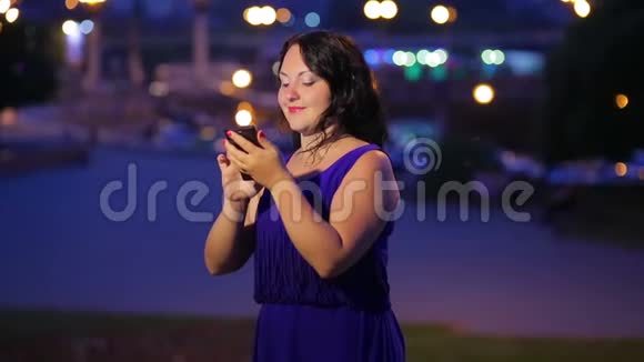 晚上一个穿着蓝色裙子的年轻女人在街上看着智能手机上的留言微笑着梦幻般的样子视频的预览图