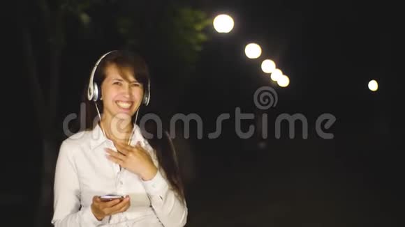 戴耳机的女人用手触摸电话在城市公园的夜晚欢笑和微笑视频的预览图