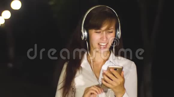 戴耳机的女孩用手在电话上交流晚上在城市公园欢笑和微笑视频的预览图