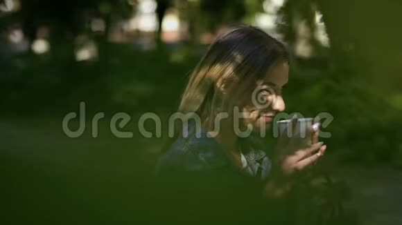 年轻的美女在夏日公园的长椅上喝咖啡放松视频的预览图