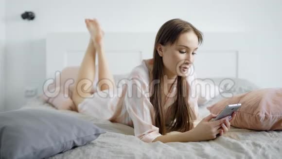 一个美丽而有趣的年轻女孩躺在床上的肖像她的空闲时间与智能手机视频的预览图