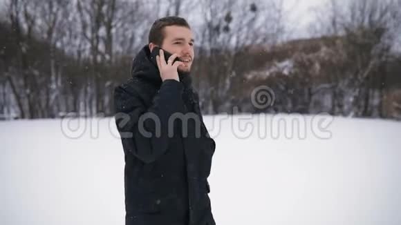 布吕内特青年肖像穿着黑色引擎盖在雪地冬季公园通过电话交谈慢动作视频的预览图