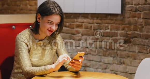 布鲁内特漂亮的年轻女性肖像穿着金色毛衣和红唇使用智能手机阁楼风格的咖啡馆背景晚上视频的预览图