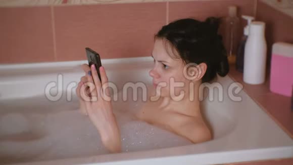 年轻漂亮的黑发女人洗澡在手机上看视频视频的预览图