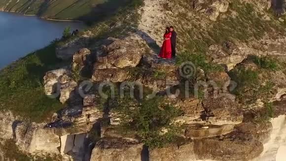 空中飞行围绕相爱的情侣站在水边的悬崖边穿着漂亮红裙子的女人穿黑色衣服的人日落视频的预览图