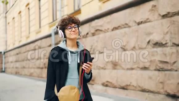 快乐的年轻女士正通过耳机听音乐在秋天独自一人在街上手持智能手机视频的预览图