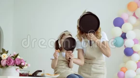 一个带着一个小女儿的漂亮女孩把脸藏在煎锅后面一个穿着围裙和妈妈在一起的小女孩视频的预览图