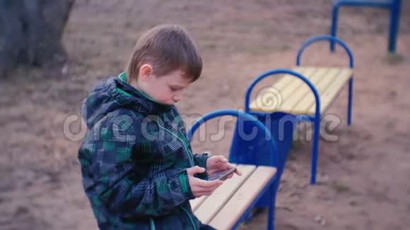 男孩坐在公园里的长凳上用手机玩游戏看着相机微笑着视频的预览图