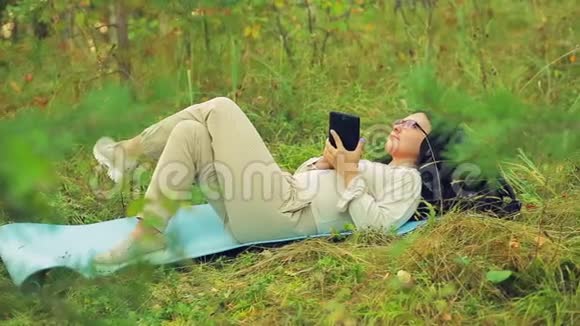 一个戴眼镜的年轻女人躺在公园的草地上在平板电脑上用信使交流视频的预览图