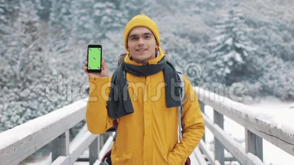 一位穿着冬装的年轻迷人男子的肖像展示了一部带有绿色屏幕的智能手机看着相机视频的预览图