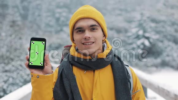 一位穿着冬装的年轻迷人男子的肖像展示了一部带有绿色屏幕的智能手机看着相机视频的预览图