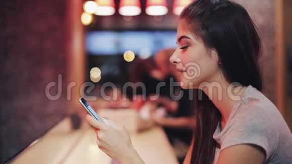 年轻女子坐在酒吧里喝着利曼德用她的智能手机旁边的霓虹灯酒吧标志享受夜生活的女人视频的预览图