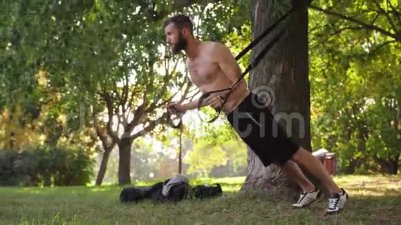 有胡子的年轻运动员在公园的一棵树附近的健身圈上做俯卧撑4K慢节奏的电影视频的预览图