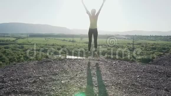 年轻的运动女孩跳起来在日落时举起她的手在山上的岩石边缘快乐女孩视频的预览图
