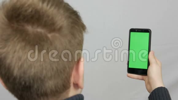 一个十几岁的男孩背对着相机站着手里拿着一部黑色智能手机上面有一个白色的绿色屏幕视频的预览图