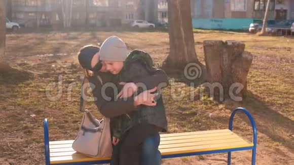 妈妈亲吻并拥抱坐在一栋多层建筑院子里的长凳上的儿子视频的预览图