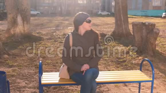 年轻漂亮的深褐色太阳镜的黑发女郎环顾四周等着有人坐在多人的城市院子里的长凳上视频的预览图