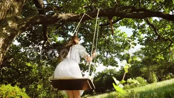 美丽的女孩留着白色连衣裙的长发在夏天的公园里荡秋千慢动作视频的预览图