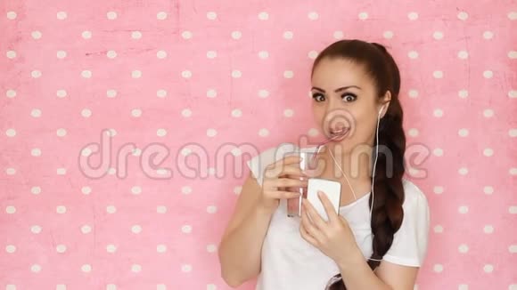 戴着耳机的年轻女人通过吸管喝水果奶昔微笑着看着他的智能手机屏幕这就是视频的预览图