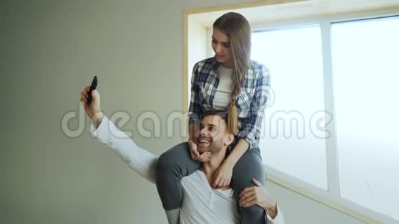 快乐的年轻夫妇在家里的厨房里玩得很开心女孩坐在男朋友脖子上而他正在拍自拍肖像视频的预览图
