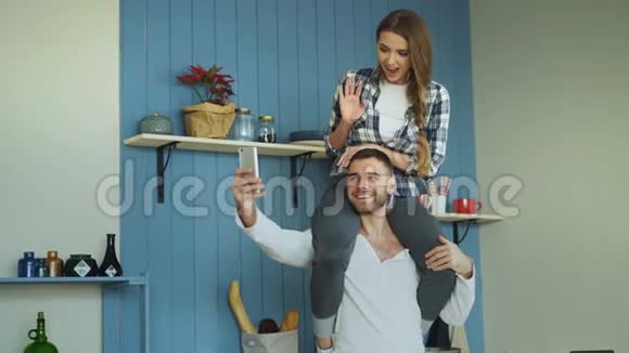 快乐的年轻夫妇在家里的厨房里玩得很开心女孩坐在男朋友脖子上而他正在拍自拍肖像视频的预览图