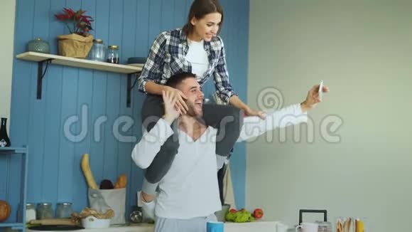 快乐的年轻夫妇在家里的厨房里玩得开心在网上聊天打视频电话女孩坐在男朋友脖子上视频的预览图