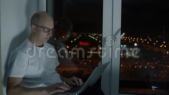 戴眼镜的中年男子在笔记本电脑上打字坐在窗台上视频的预览图